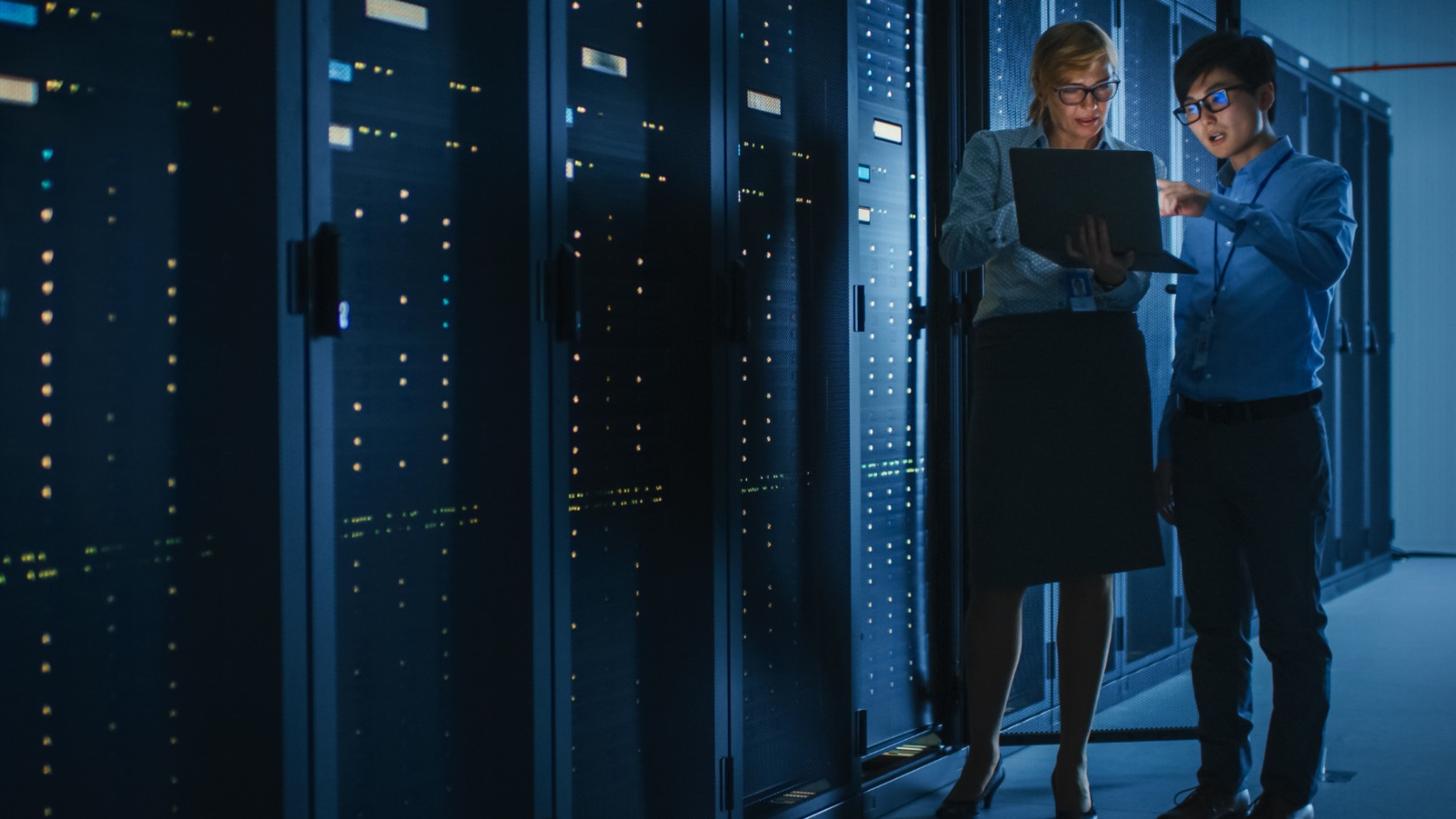 Two IT professionals reviewing information on a laptop between server racks in a data centre