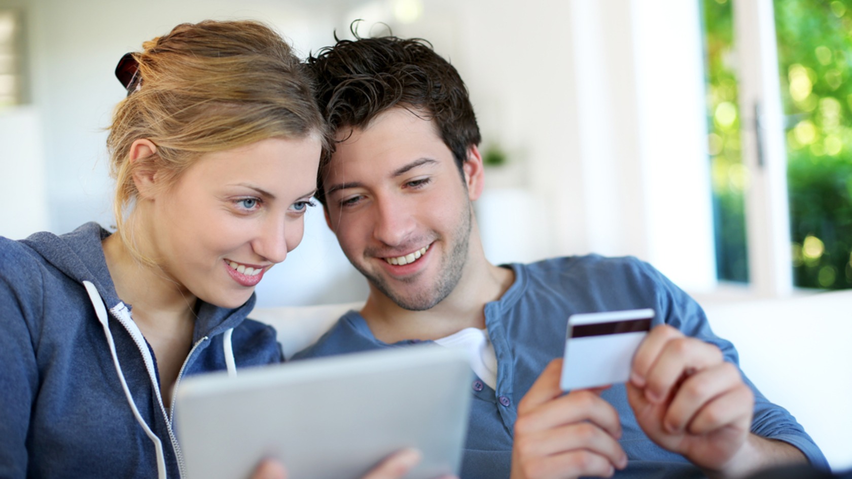 Young couple smiling at a tablet while holding a payment card to shop online, illustrating why ecommerce businesses must secure customer transactions.