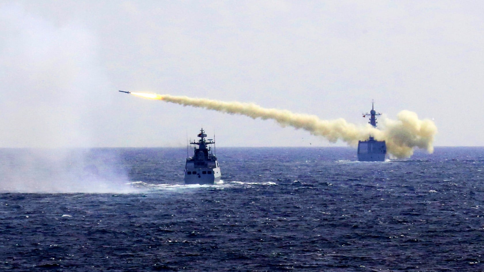 Naval warships at sea during a live fire drill, with a missile streaking across the sky trailing smoke